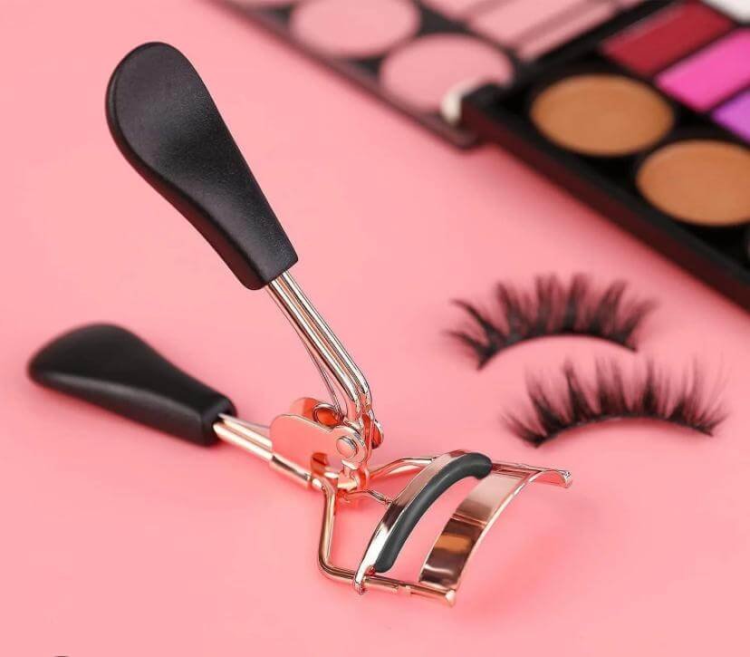 "Rose gold beauty product.An eyelash curler with black handles next to false lashes on a pink background"